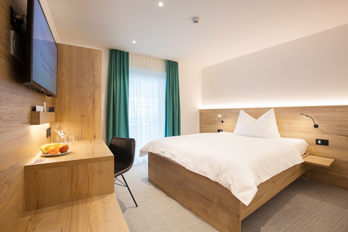 Une chambre d’hôtel moderne avec un lit en bois, un bureau avec une chaise, un téléviseur à écran plat et une grande fenêtre avec des rideaux.