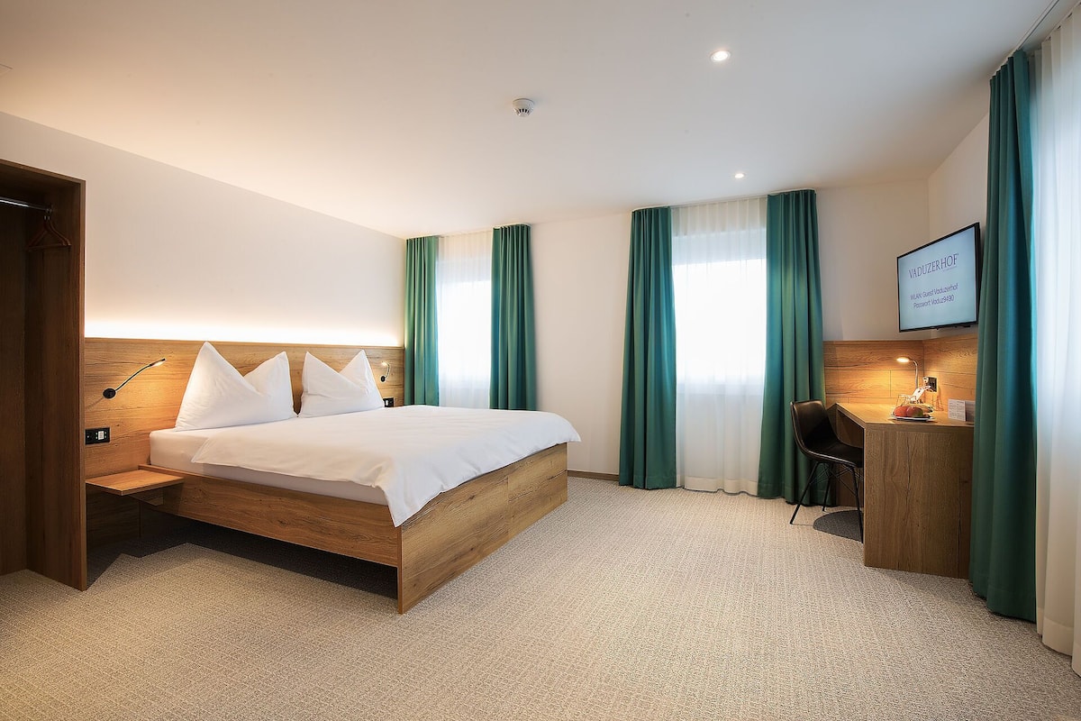 Une chambre d’hôtel avec un lit en bois, des rideaux verts, un bureau avec une chaise et un téléviseur fixé au mur qui affiche des informations sur la chambre.