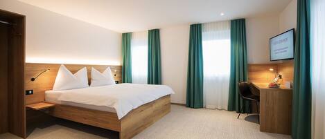 Habitación de hotel con cama de madera, cortinas verdes, escritorio con silla y televisor montado mostrando información de la habitación.