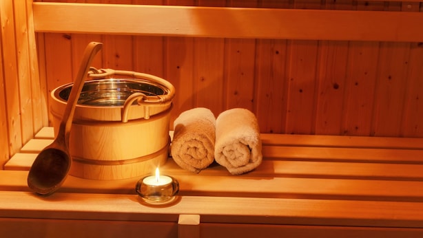 Una sauna de madera con una bañera grande de madera, una cuchara de madera, dos toallas enrolladas y una vela encendida.