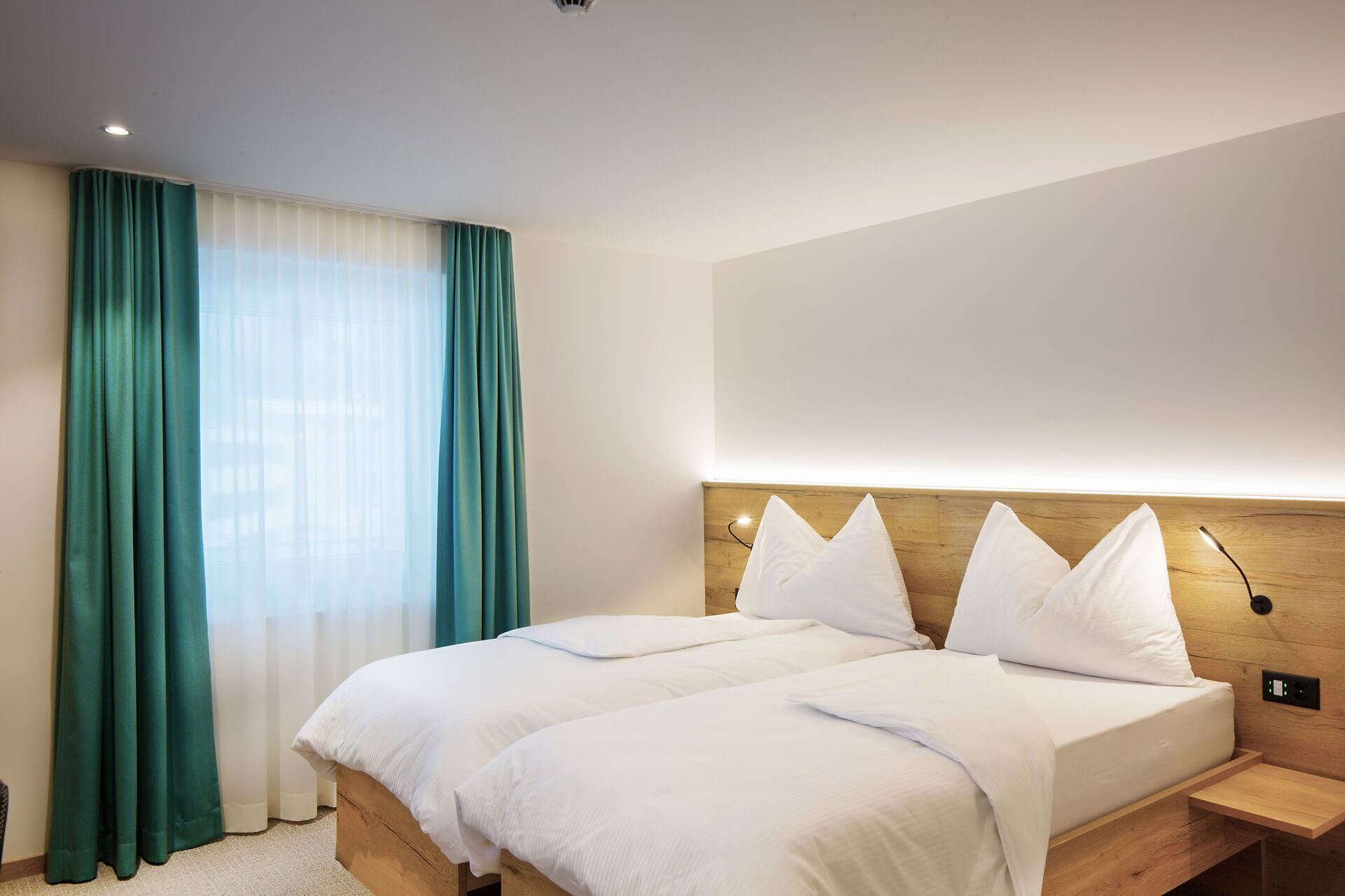 A hotel room with a double bed, a wooden headboard, a bedside table, and a large window with curtains.