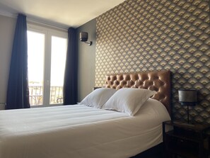 A hotel room with a large bed, a headboard, a wall with a patterned wallpaper, a window with curtains, and a nightstand with a lamp. - Hotel Regina (Sète)