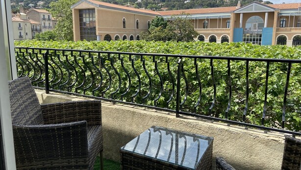 A balcony with wicker furniture and a view of a building with a red roof and arched windows. - Hotel Regina (Sète)