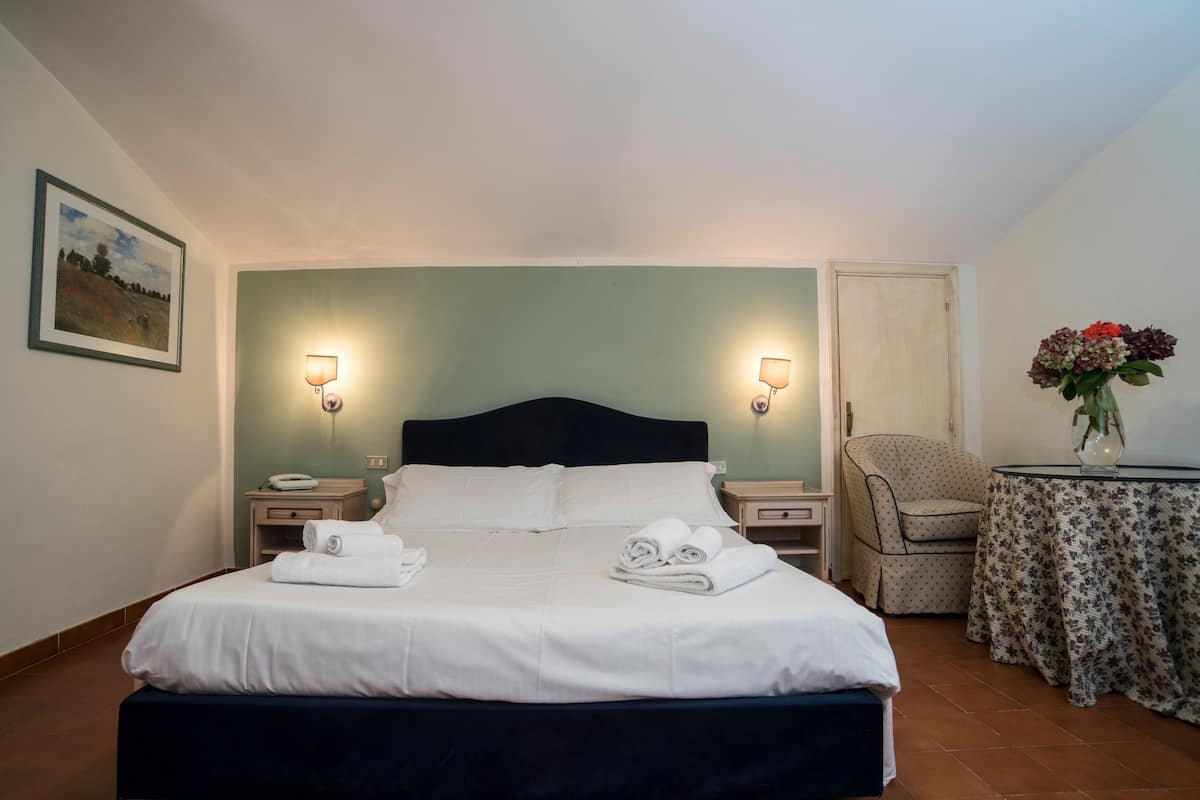 A neatly made bed with white linens and towels, two bedside lamps, a chair, a small table with a floral tablecloth, and a framed picture on the wall.