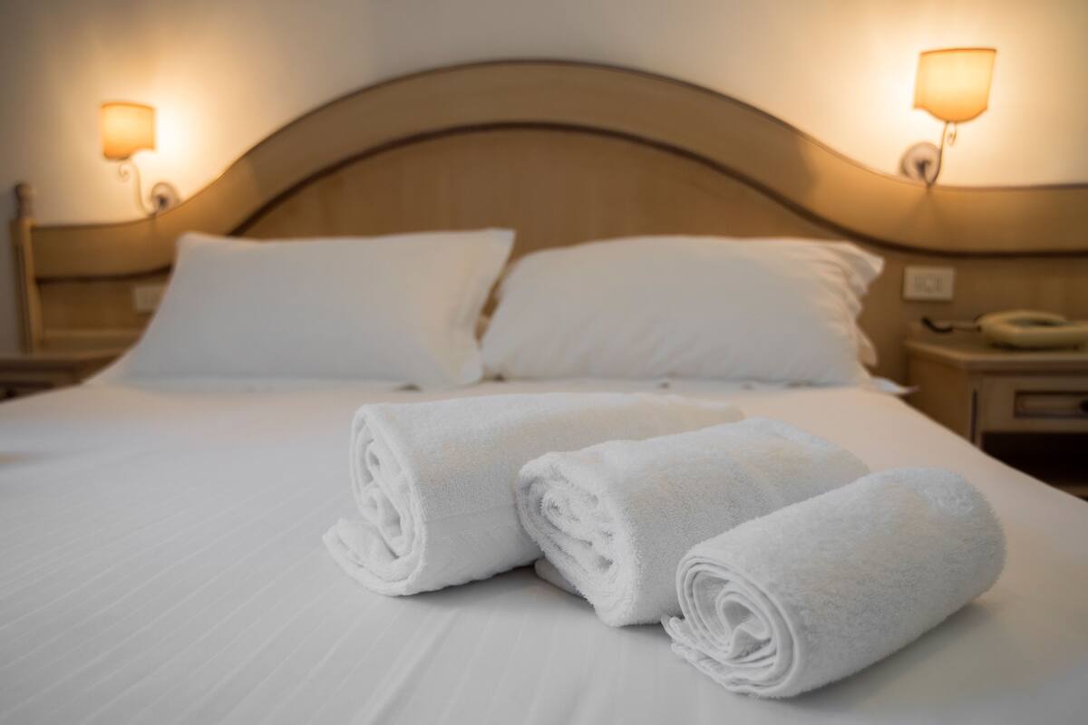 A neatly made bed with white linens and two rolled towels on top. There are two bedside lamps on either side of the bed.