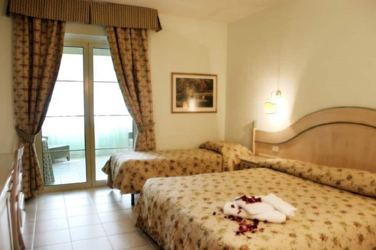 A hotel room with two beds, a sliding glass door leading to a balcony, a framed picture on the wall, and a bedside lamp.