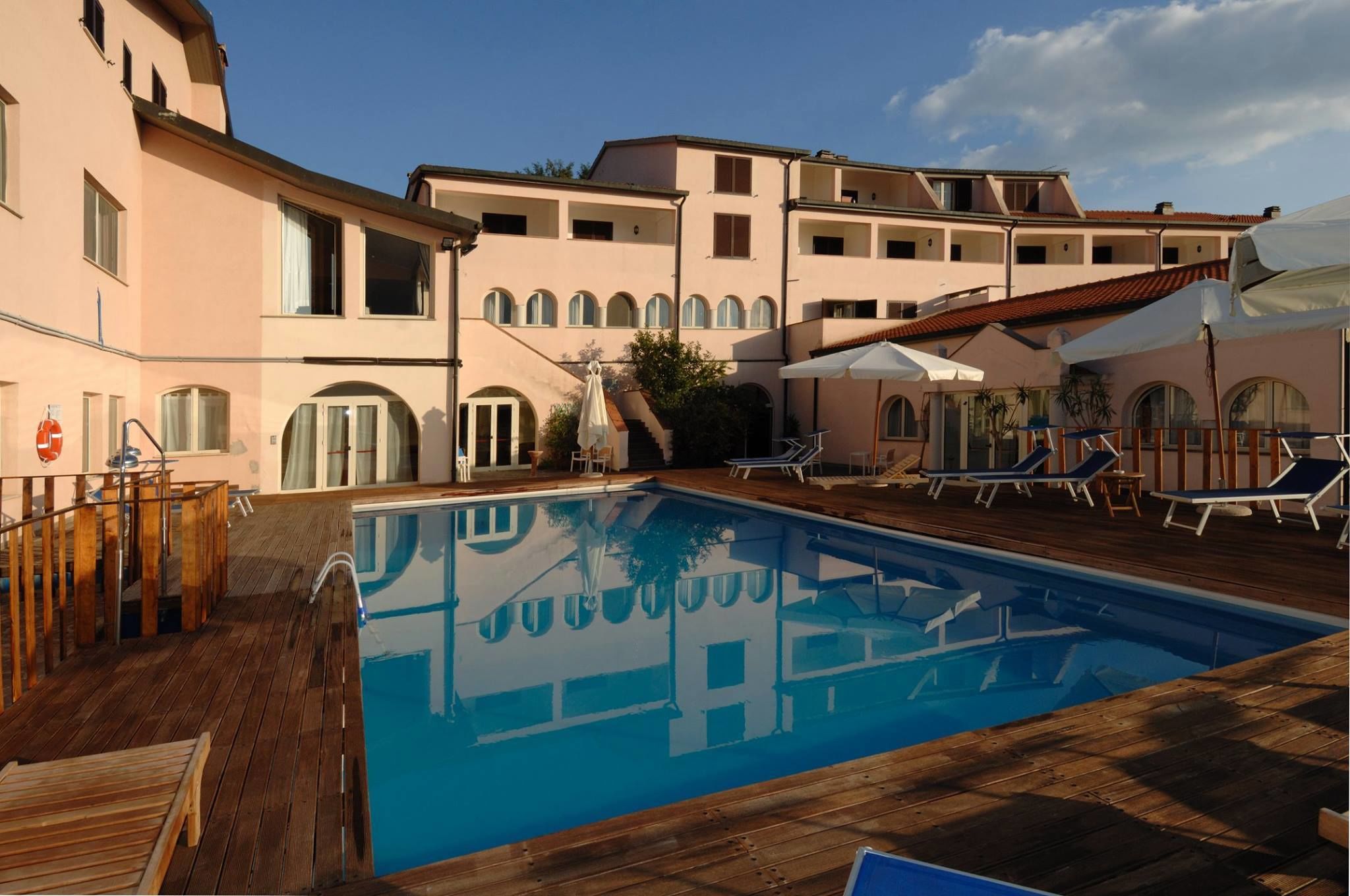 A pool area with wooden deck, lounge chairs, and a building with arched windows and balconies.