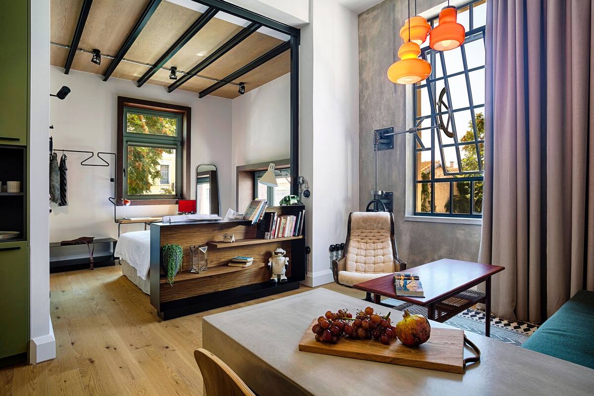 Quarto moderno com cama, escrivaninha, cadeira e mesa de jantar. Há uma janela com cortinas, uma estante de livros e um vaso de planta sobre a mesa.