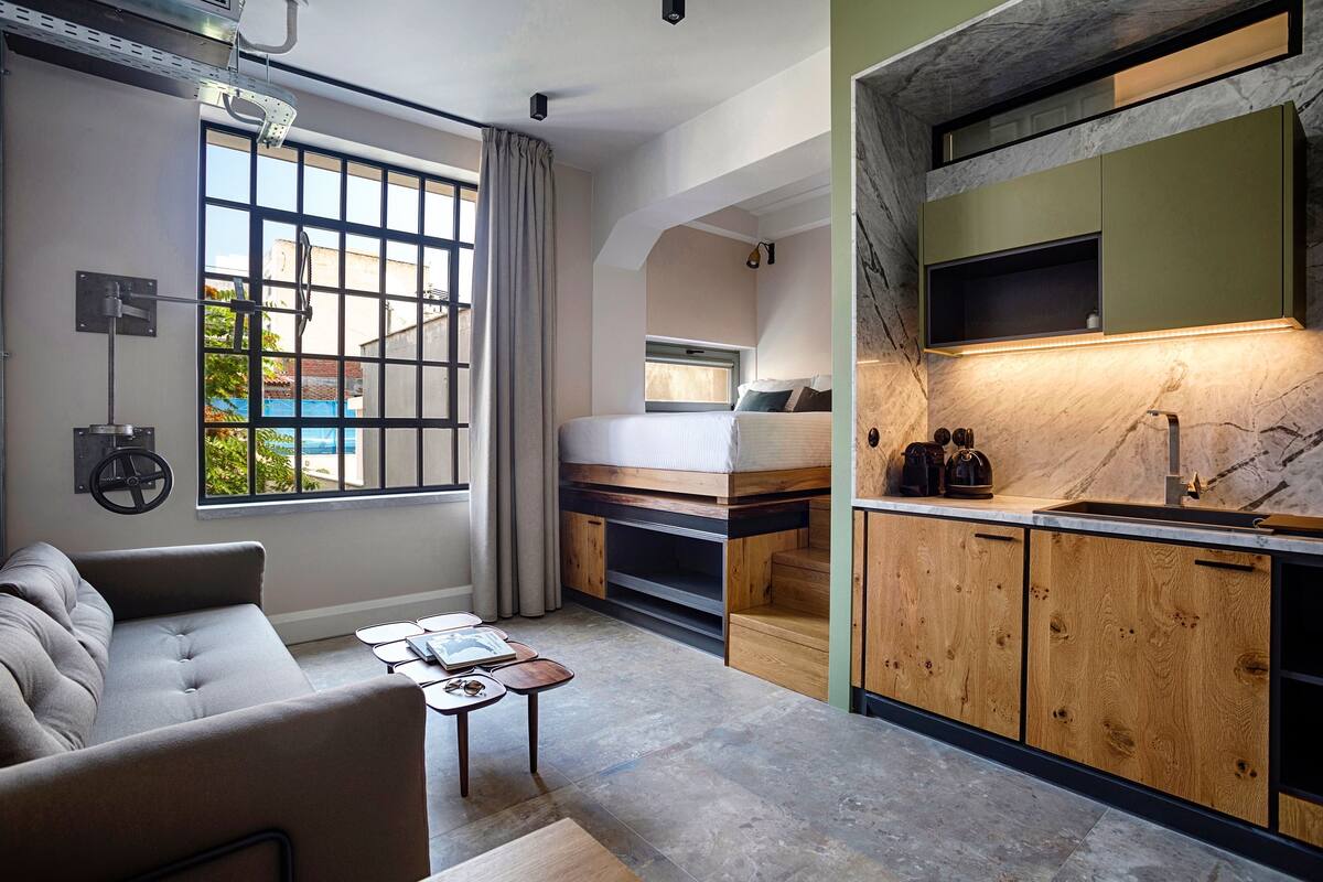 Quarto de hotel compacto com sofá, uma mesa pequena, beliche e área de cozinha com armários de madeira e bancada de mármore.