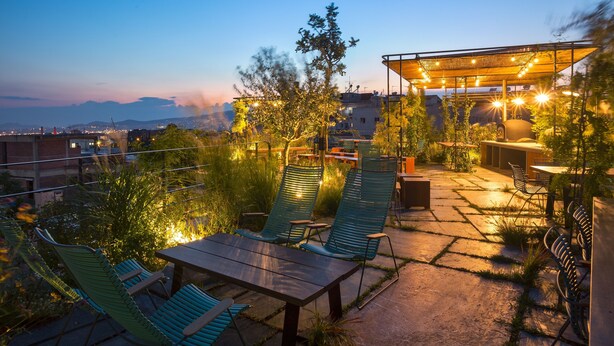 Un patio al aire libre con mobiliario y una zona de comedor cubierta, rodeado de vegetación y vistas a la ciudad.