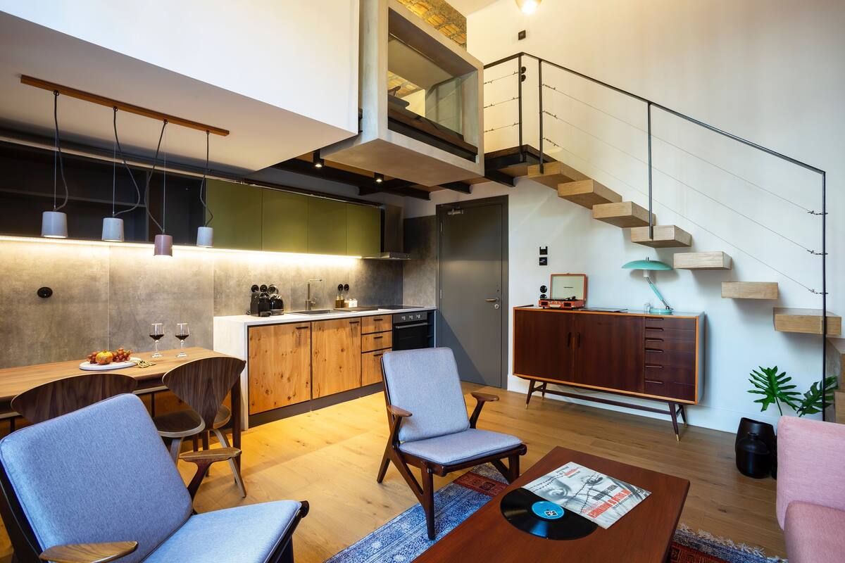Uma sala de estar moderna com uma mesa de centro de madeira, duas poltronas, uma área de jantar com mesa e cadeiras de madeira, uma cozinha com forno embutido e pia, e uma escada com corrimão de vidro.