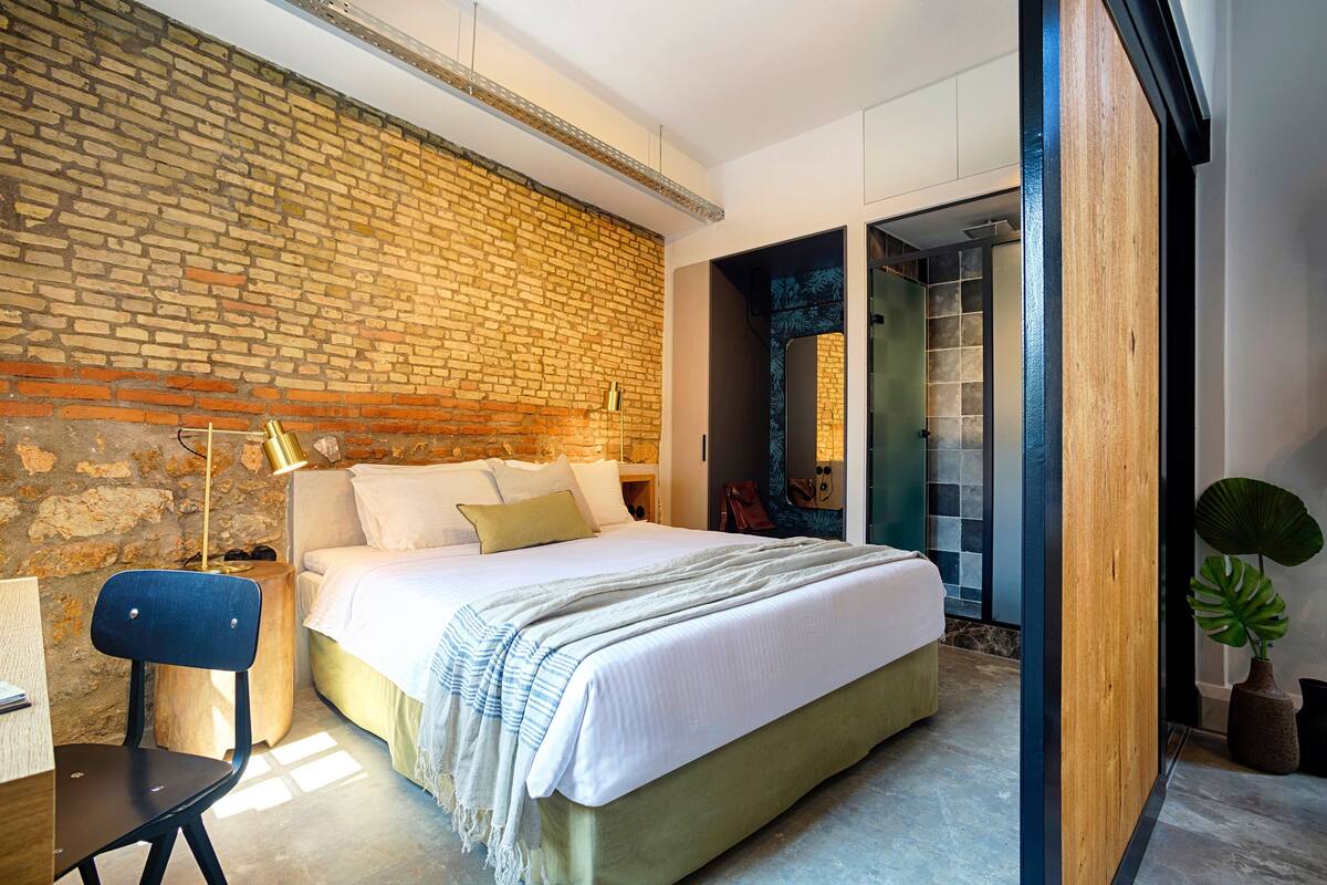Quarto moderno com parede de tijolos, cama com roupa de cama branca, mesa de cabeceira de madeira, escrivaninha com cadeira e uma planta em vaso.