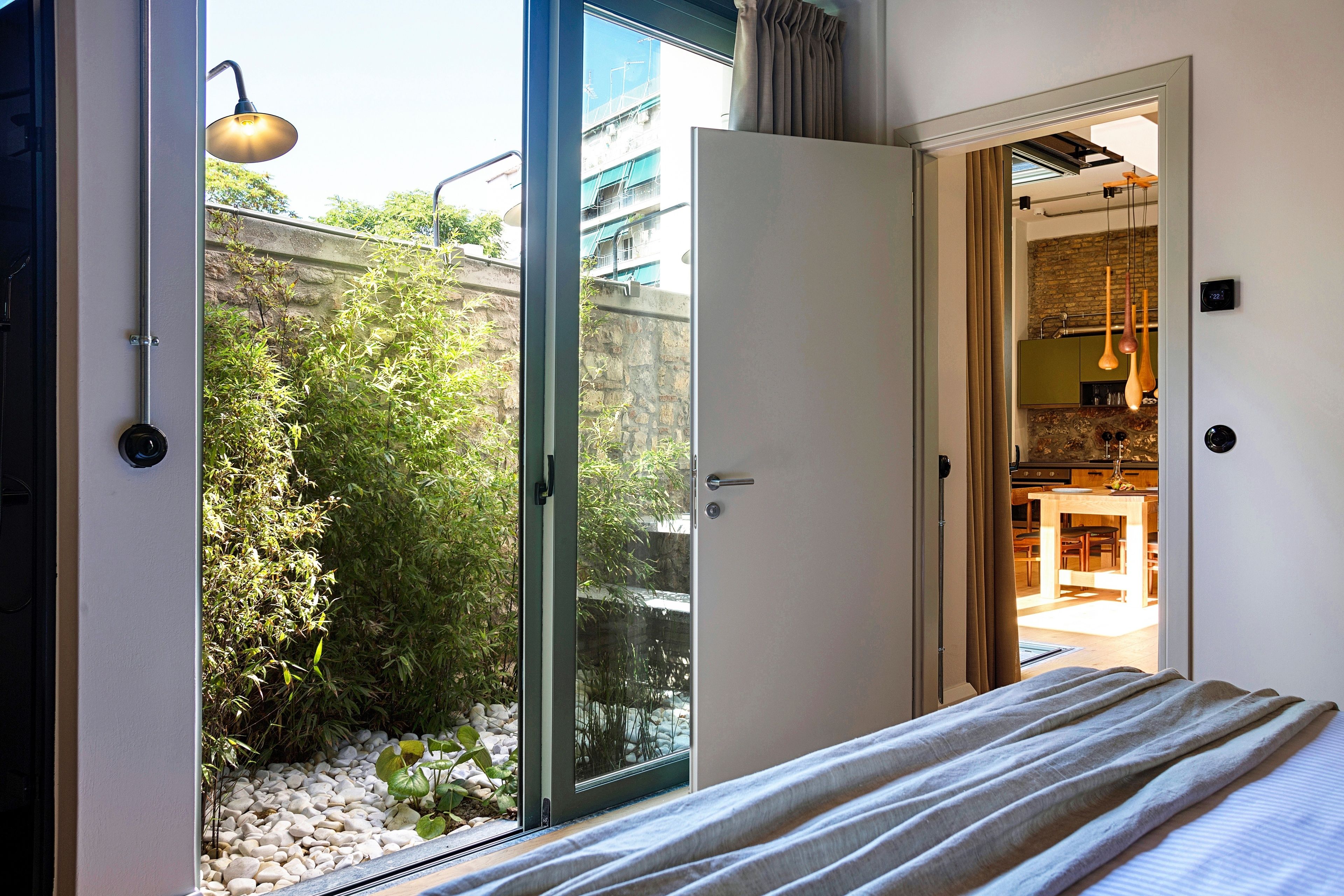 A bedroom with a bed, a sliding glass door leading to a balcony with plants and a view of buildings.