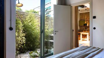 A bedroom with a bed, a sliding glass door leading to a balcony with plants and a view of buildings.