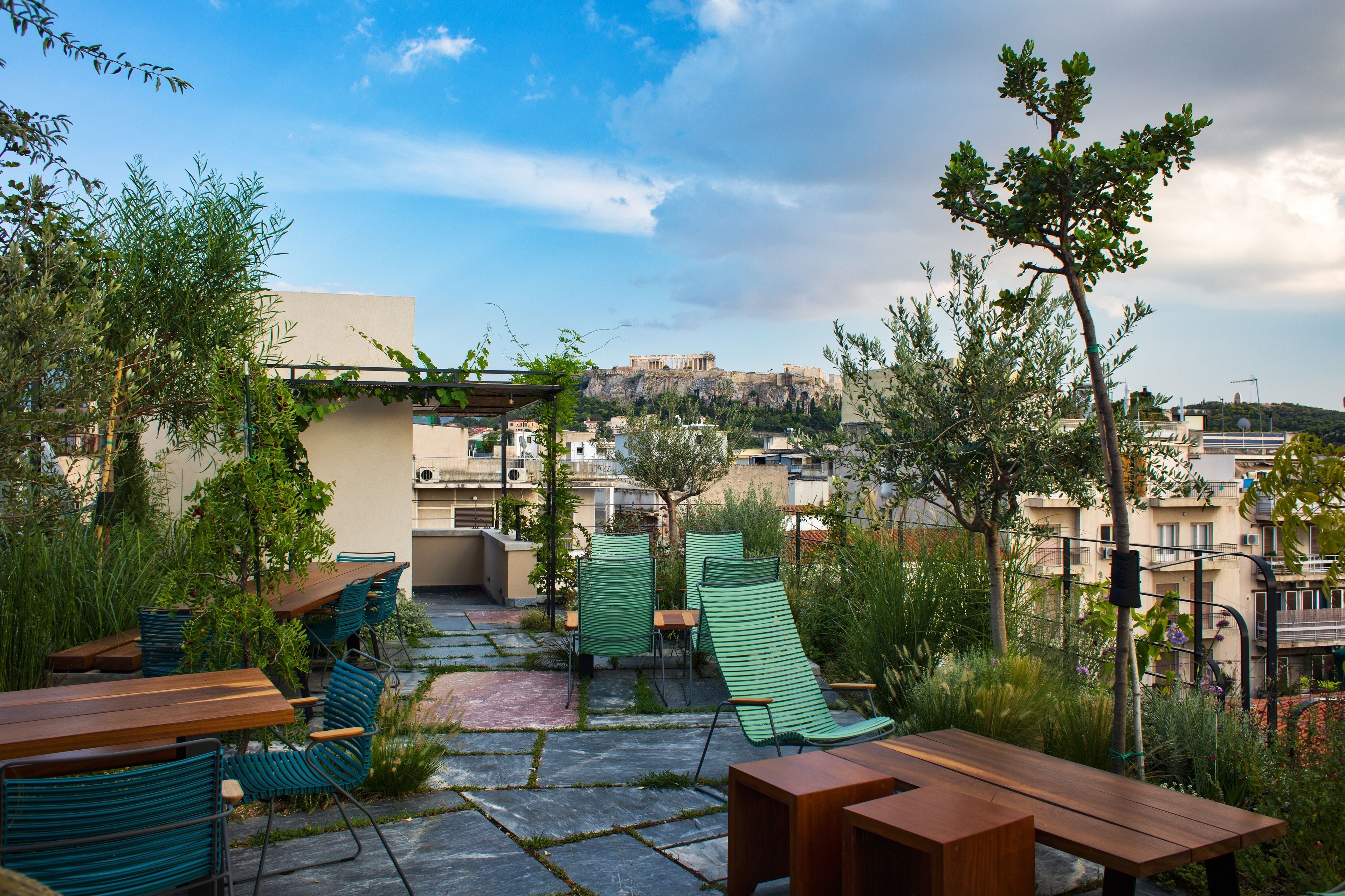 Eine Außenpatio mit Holztischen und -stühlen, umgeben von Grünpflanzen und mit Blick auf eine Stadtlandschaft mit Gebäuden und einer Erhebung im Hintergrund.