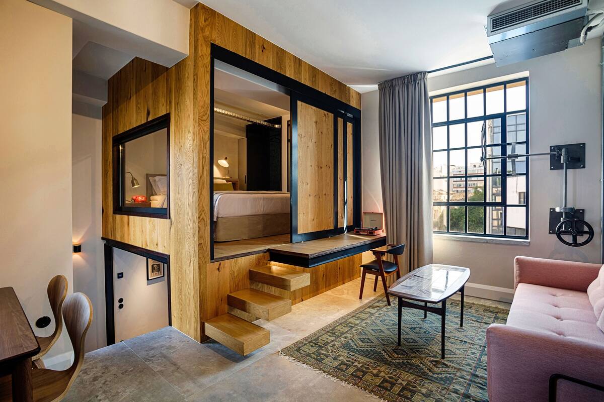 Quarto de hotel moderno com escada de madeira, espelho grande, sofá, mesa pequena e vista da cidade por grandes janelas.