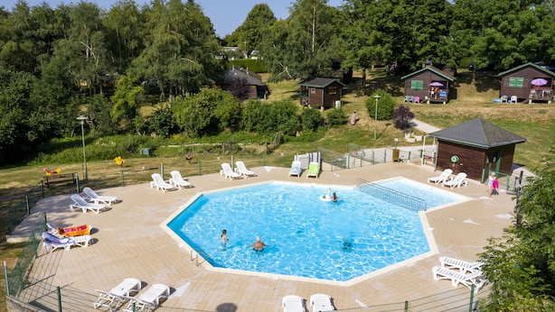 Una piscina con sillas de descanso y un área cercada, rodeada de árboles y pequeñas cabañas.