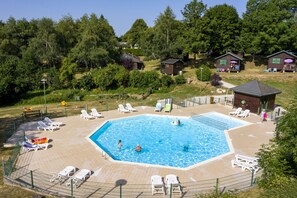 Una piscina con sillas de descanso y un área cercada, rodeada de árboles y pequeñas cabañas.