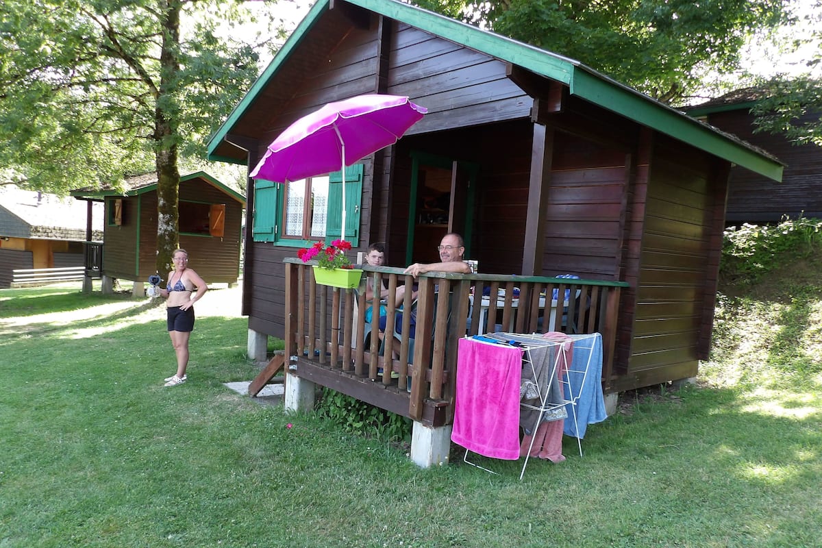 Een houten chalet met een veranda, een tafel met een roze parasol en een groene bloempot met bloemen.