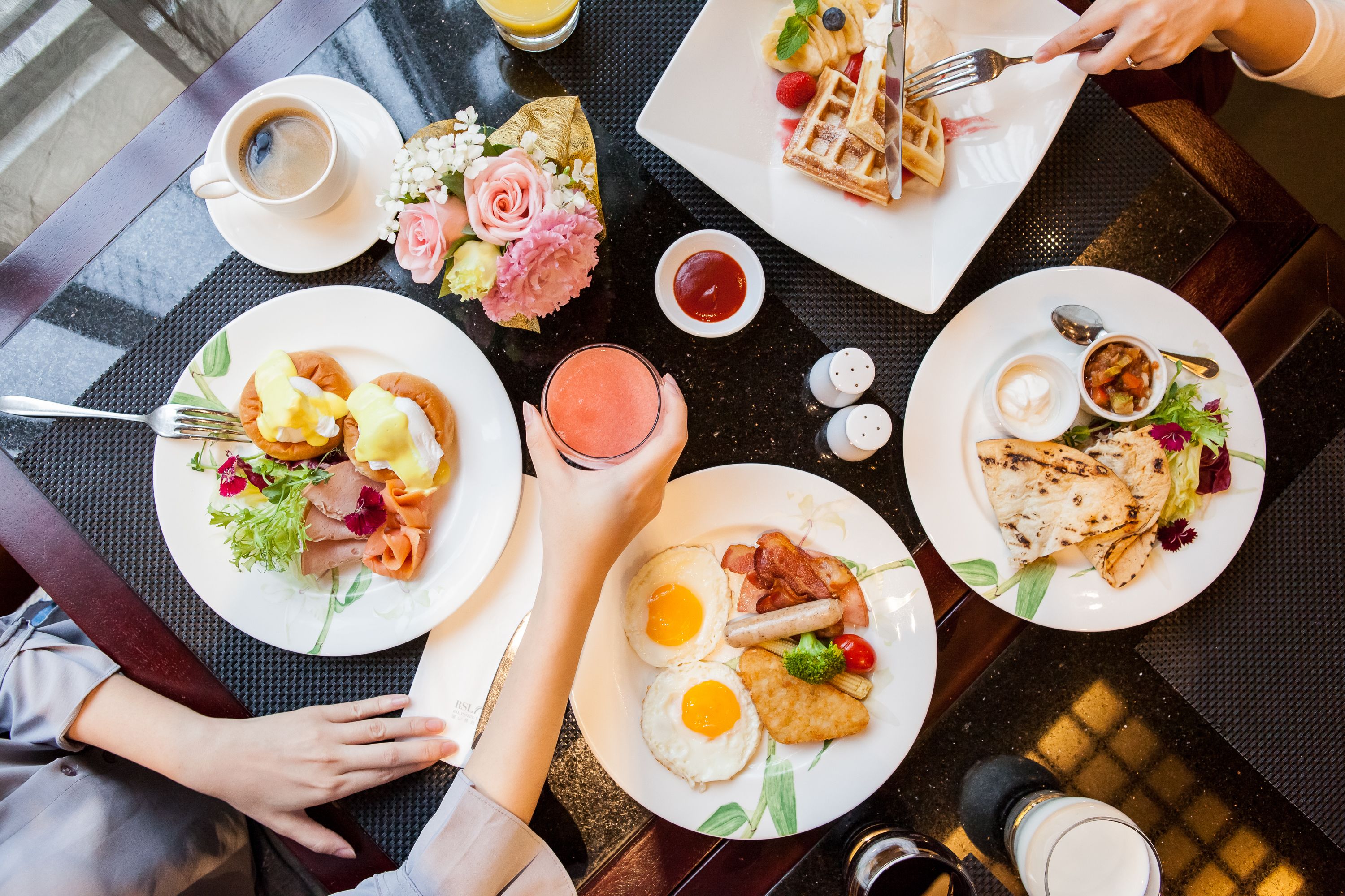 A breakfast setting with multiple plates of food, including waffles, eggs, and toast, along with beverages and a vase of flowers.