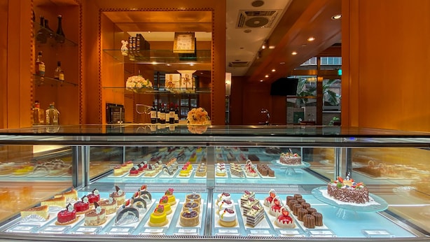 Een bakkerijdisplaykast met diverse desserts en een aantal flessen op planken in de achtergrond.
