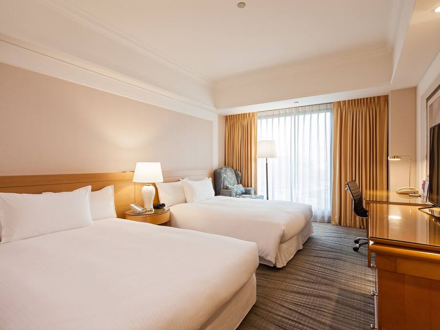 A hotel room with two beds, a desk, a chair, a TV, and a window with curtains.