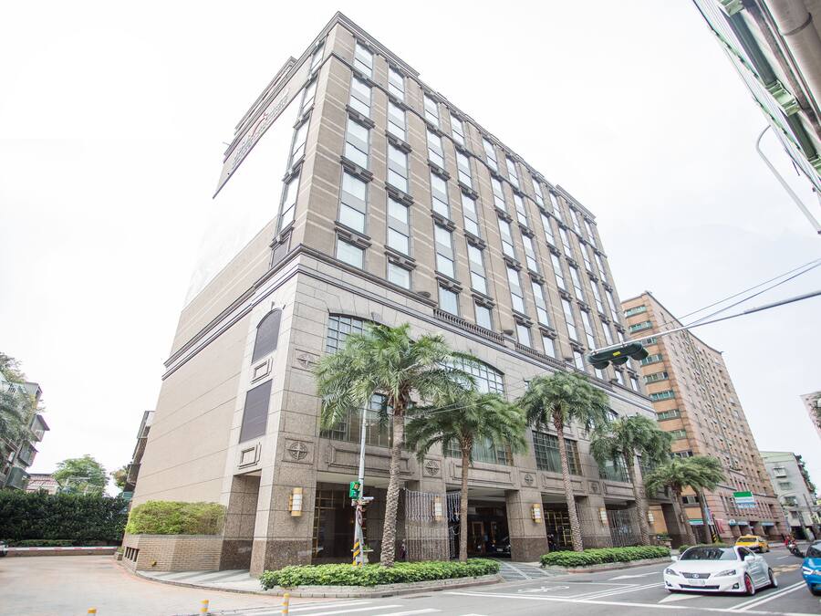 Un hotel de varios pisos con fachada moderna, palmeras en primer plano y vista a la calle con coches.