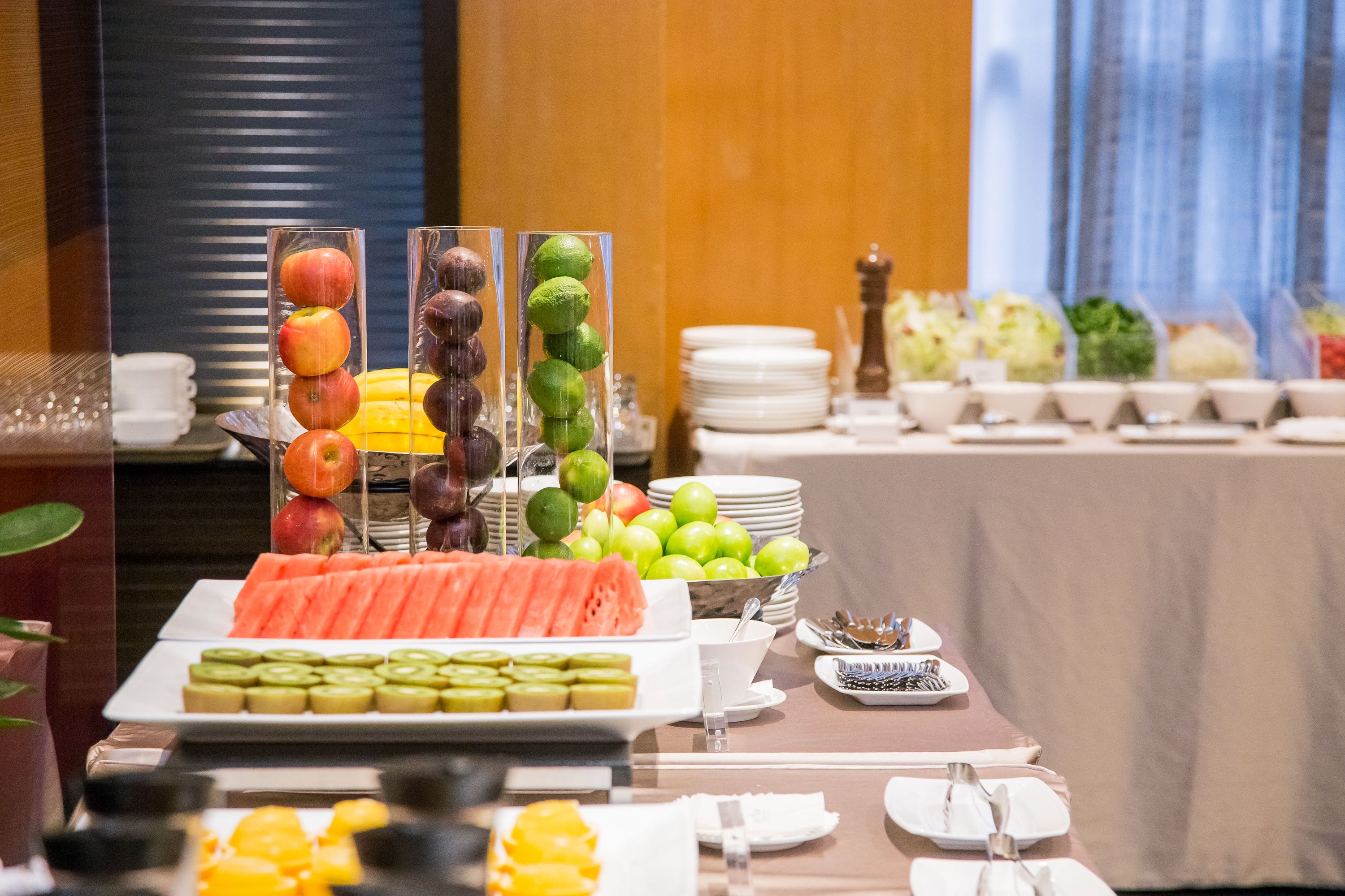 A buffet with various fruits and vegetables, including apples, limes, and watermelon, arranged on a table with plates and cutlery.
