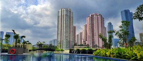 A swimming pool with a bridge, surrounded by greenery and tall buildings in the background.