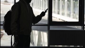 Una persona de pie en una habitación con amplios ventanales, sosteniendo un dispositivo móvil.
