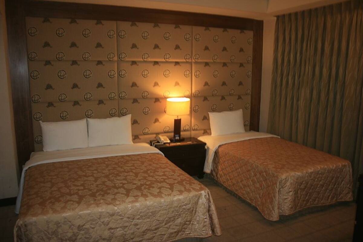 A hotel room with two beds, a headboard with a patterned design, a nightstand with a lamp, and a curtain on the window.