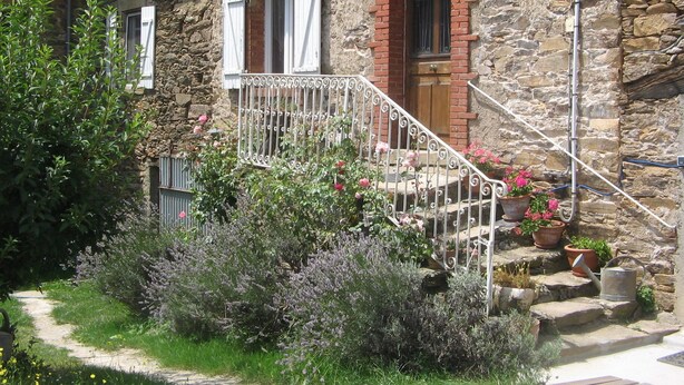 Un edificio de piedra con puerta de madera, contraventanas blancas y una escalera de metal que lleva a un balcón.