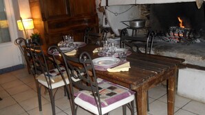 Una mesa de comedor de madera dispuesta con platos, vasos y cubiertos. Sillas con asientos acolchados rodean la mesa. Se ve una chimenea con fuego al fondo.