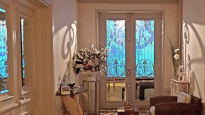 A room with a large window with decorative iron framing, a potted plant, a small table, and a wicker chair.