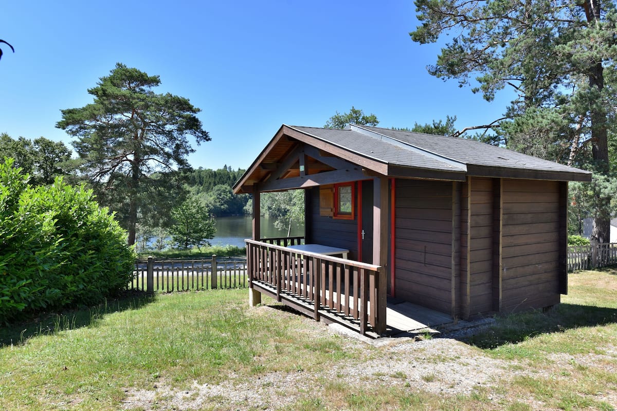 Une petite cabane en bois dotée d’un porche, entourée de verdure et bordée par un lac.