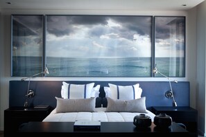A bedroom with a large window, a bed with white bedding, two bedside tables with lamps, and a view of the sea. - North (Cape Town)