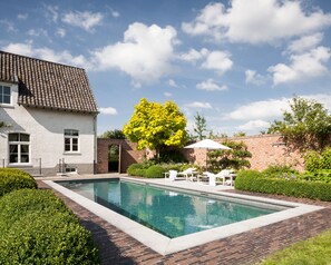 A rectangular swimming pool with a brick border, surrounded by a well-maintained garden and a white building with a tiled roof. - B&B De Lievde (Lievegem)