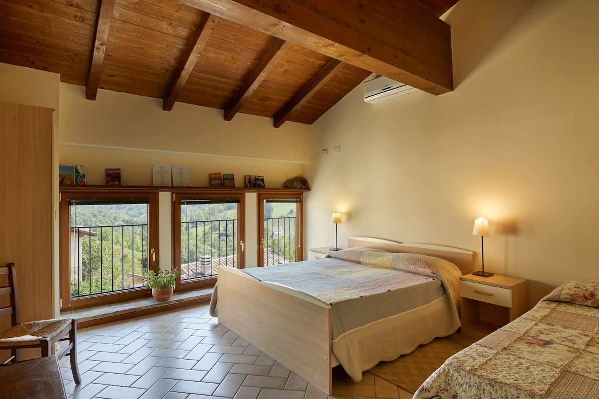 A bedroom with a wooden ceiling, a bed with a quilt, a nightstand with a lamp, and a view of a balcony with a railing.