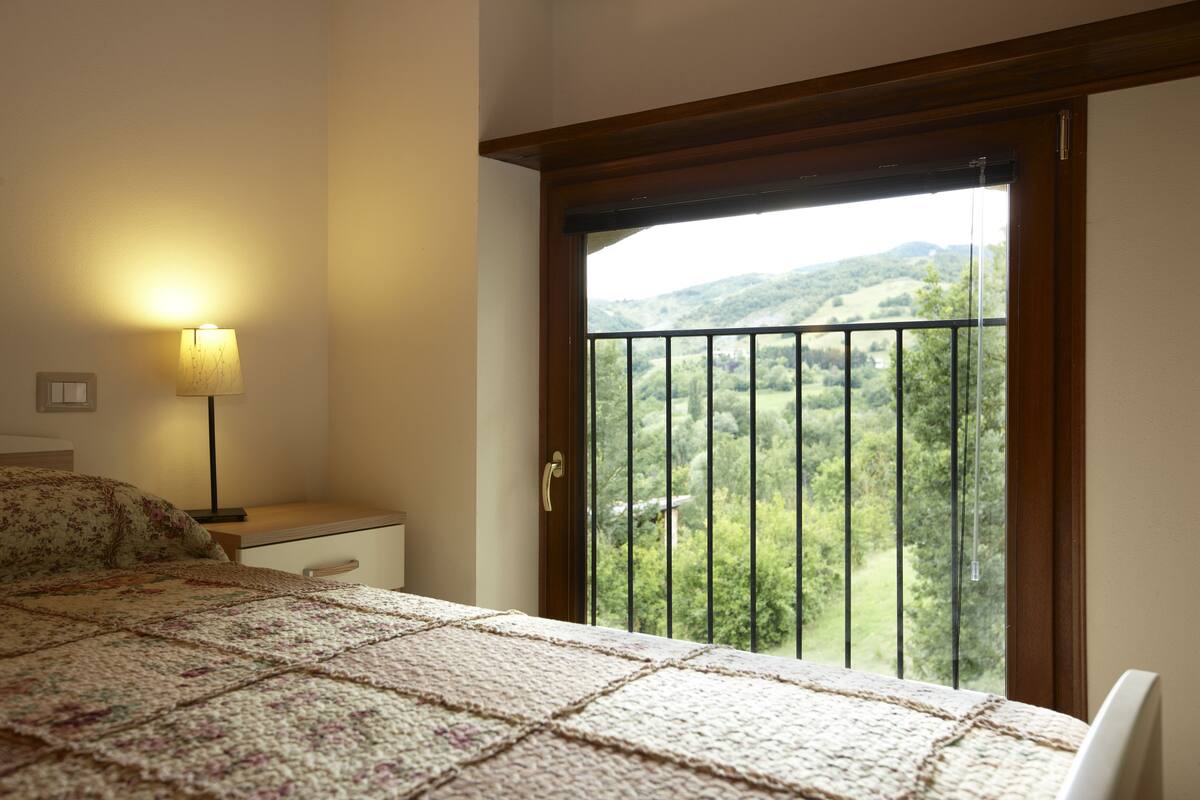 A bedroom with a bed, a nightstand, a lamp, and a window with a view of a green landscape.