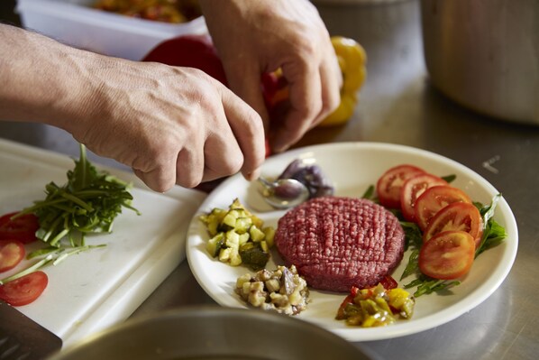 Una persona preparando una comida con un plato que contiene tomates en rodajas, lechuga y una hamburguesa.