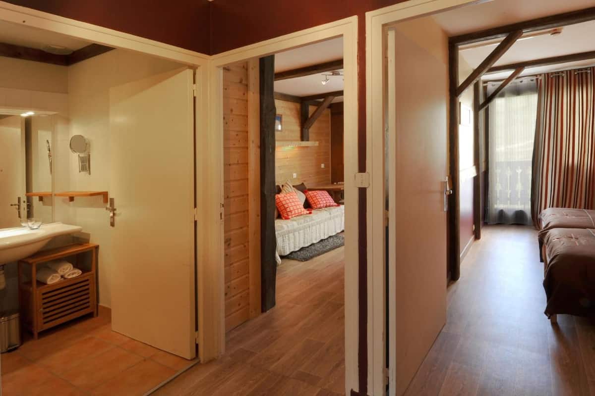 Habitación de hotel con piso de madera, cama con ropa de cama roja y blanca, baño con lavamanos y espejo, y vista de la habitación a través de una puerta abierta.
