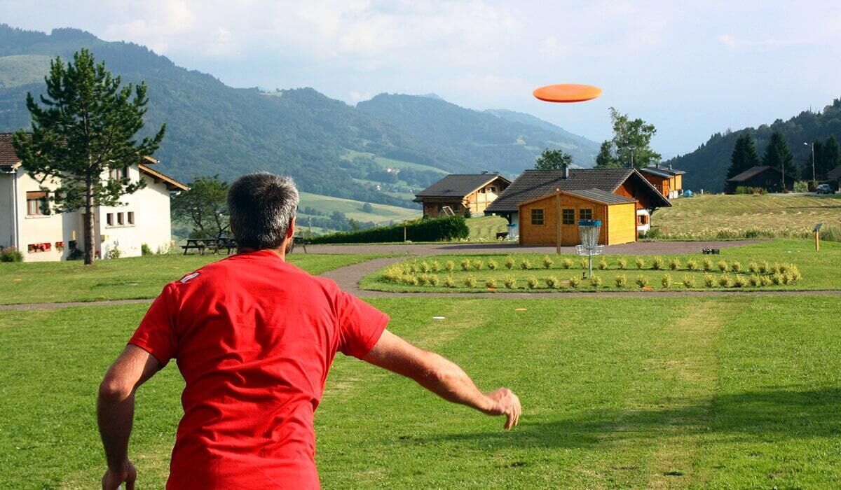A man in a red shirt throwing a frisbee in a grassy field with houses and mountains in the background.