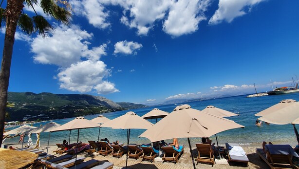 A beach with sun loungers and umbrellas, clear blue water, and a boat in the distance. - Onar Corfu Aparts & Studios (Corfu)