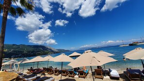 A beach with sun loungers and umbrellas, clear blue water, and a boat in the distance. - Onar Corfu Aparts & Studios (Corfu)
