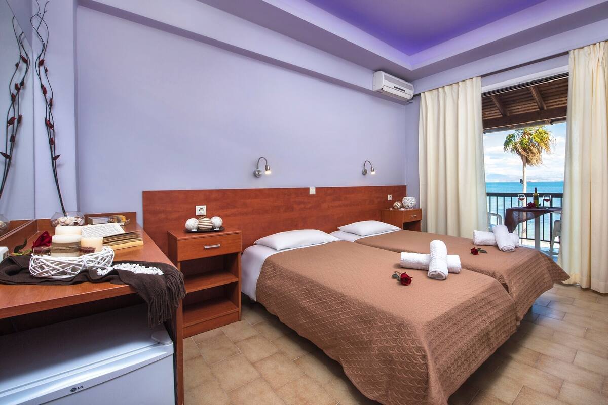 A hotel room with two beds, a wooden desk, a nightstand, a balcony with a view of the sea, and an air conditioning unit.