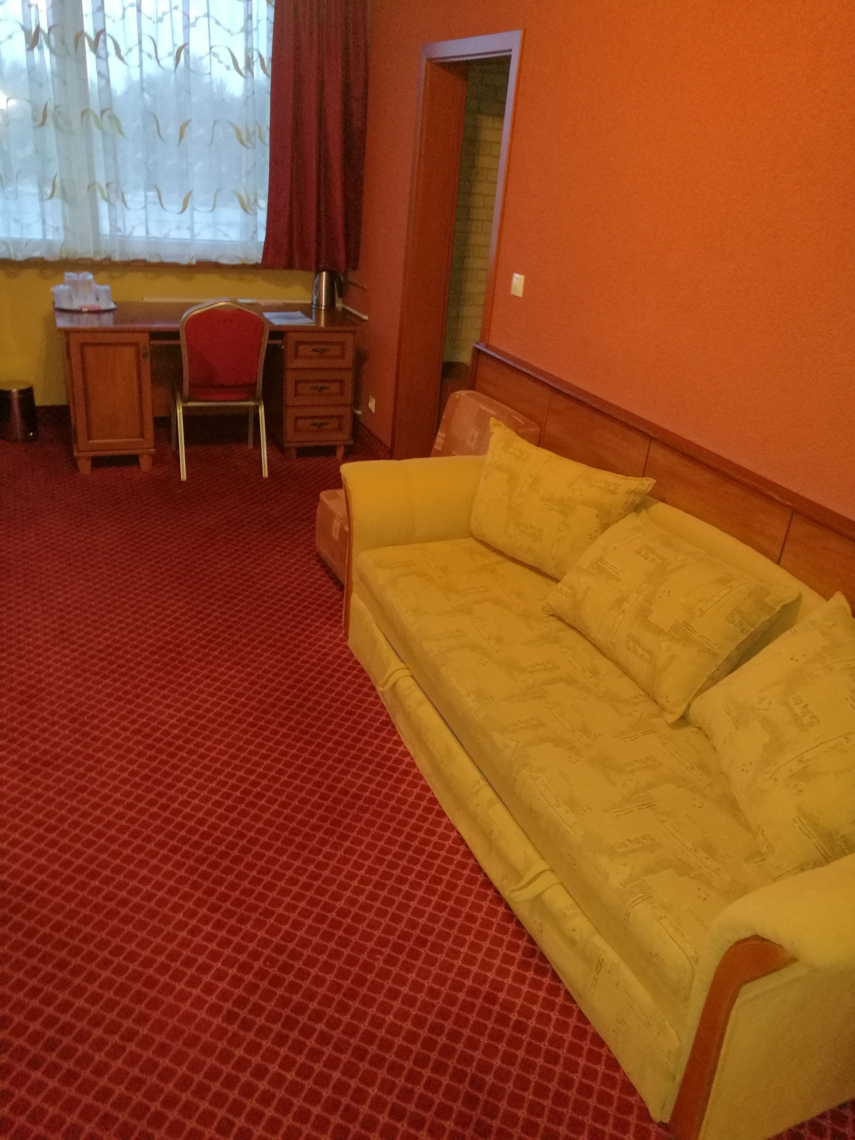 Una habitación con alfombra roja, sofá amarillo, escritorio de madera, una silla y una ventana con cortinas.