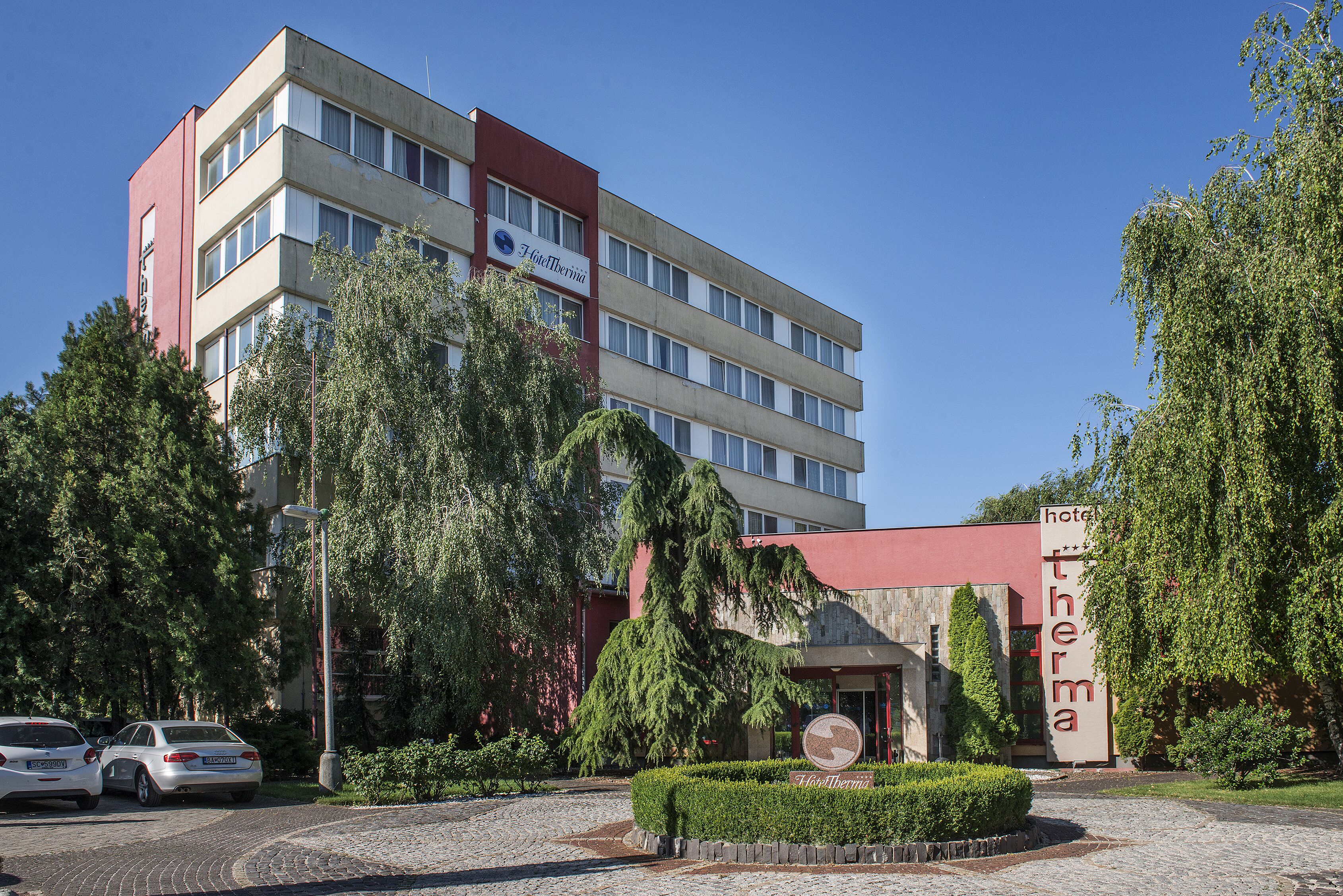 En hotels bygning med en rød og beige facade, mange vinduer og et skilt der angiver, at det er et hotel. Der er træer og et belagt område foran.