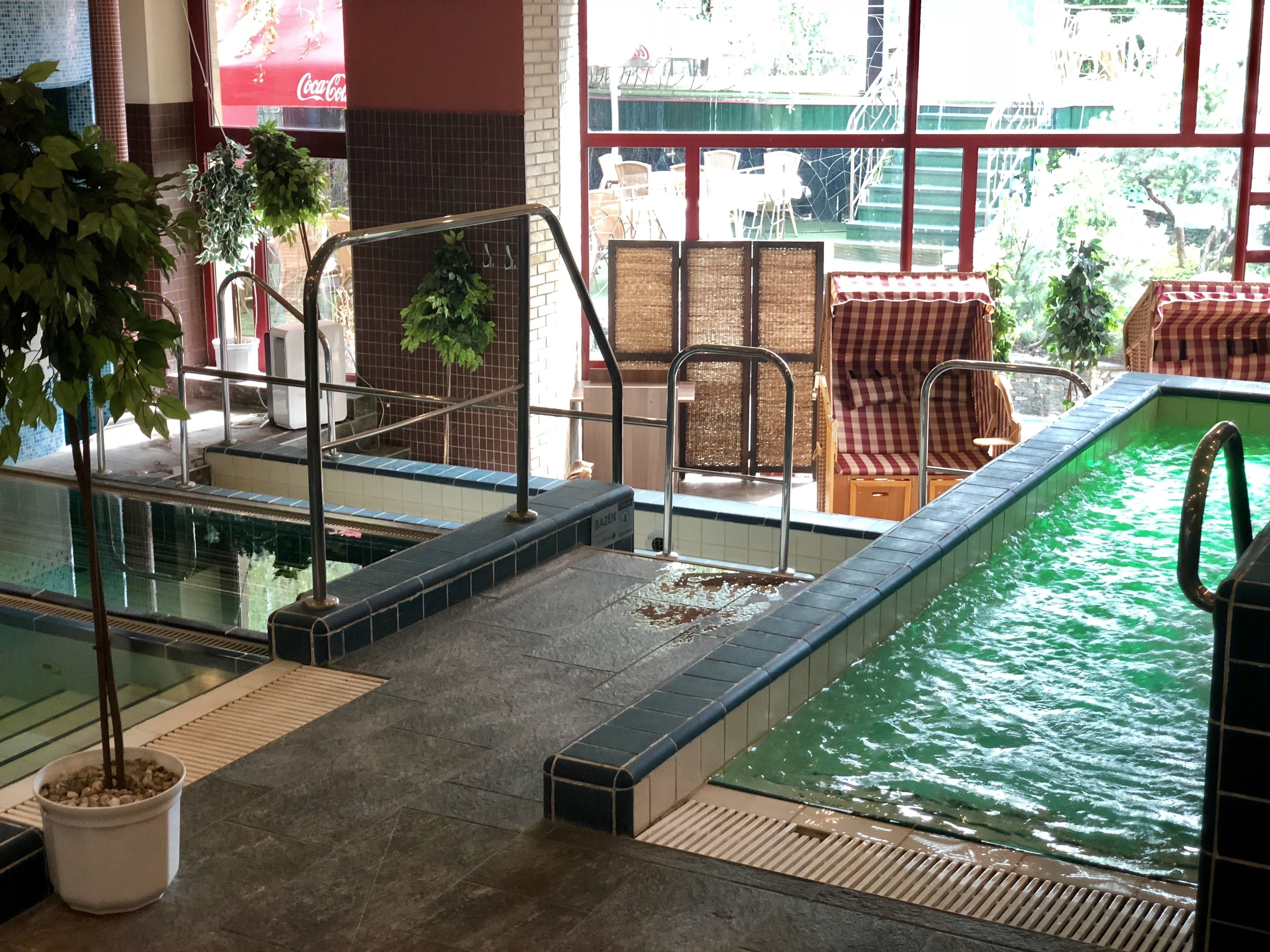 Uma piscina interna com piso de azulejos, um anúncio da Coca-Cola e cadeiras de vime perto da janela.
