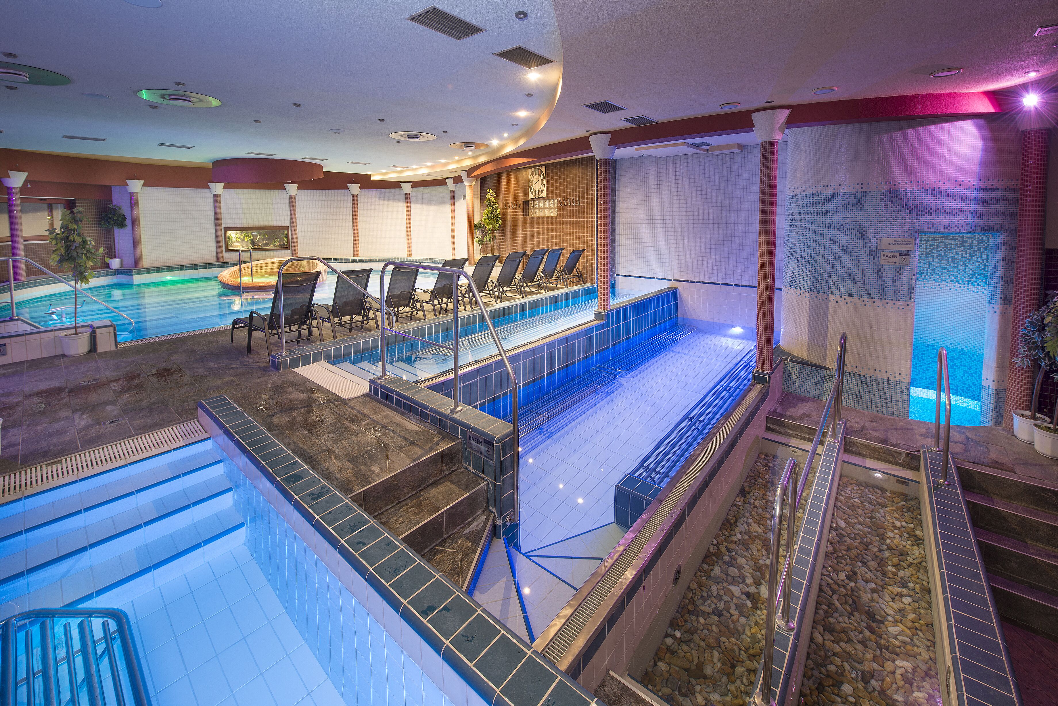 Un espace piscine d’hôtel comprenant plusieurs piscines, une terrasse ensoleillée et un coin salon avec chaises et tables.