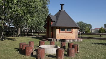 Una cabaña de madera pequeña con techo a dos aguas y chimenea, rodeada por un fogón circular y postes de madera.